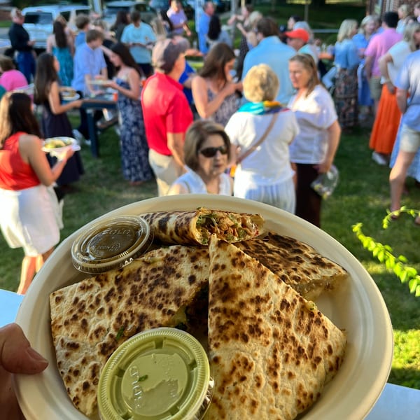 A plate of quesadillas with dipping sauces, held in front of a blurred outdoor gathering.